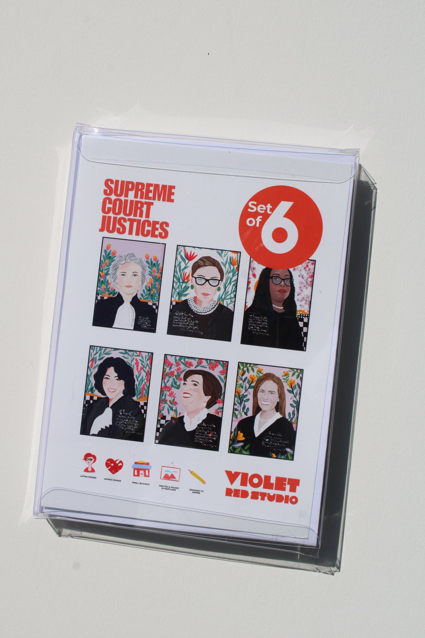 A clear plastic sleeve encloses a printed card or booklet titled 'Supreme Court Justices Set of 6' with six illustrated portraits and 'Violet Red Studio' branding, photographed on a plain light background.