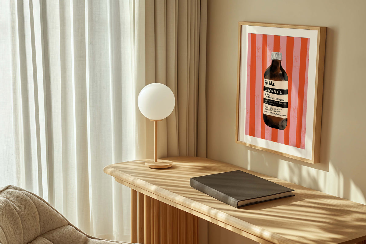 Framed artwork of a bottle on a wall above a wooden desk with a lamp and notebook.