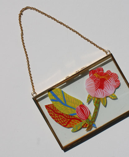 A rectangular gold-tone metal clutch bag with a clear top panel, featuring a colorful floral illustration of pink and red flowers with yellow and green leaves. A gold-tone chain is attached at the top corners, set against a plain light gray background.