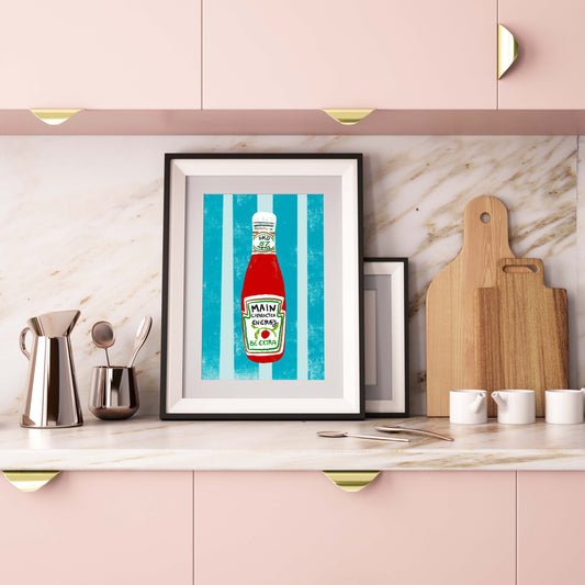 Framed artwork of a ketchup bottle on a kitchen counter with pink cabinets and marble backsplash.