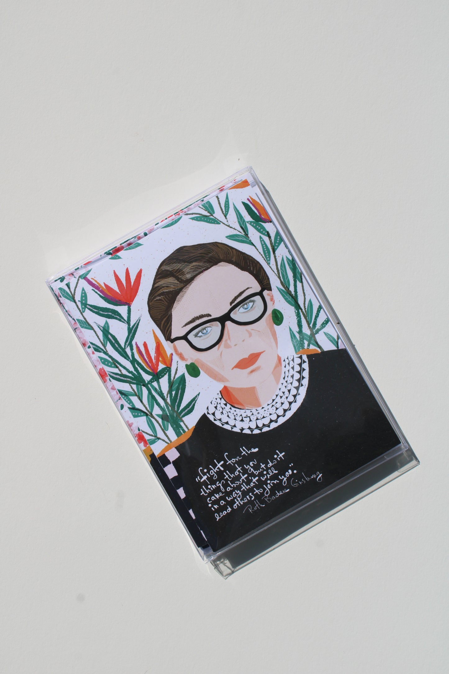 A clear plastic cassette tape case featuring a stylized portrait of Ruth Bader Ginsburg, surrounded by colorful tropical floral illustrations and handwritten text, shown on a light gray background.