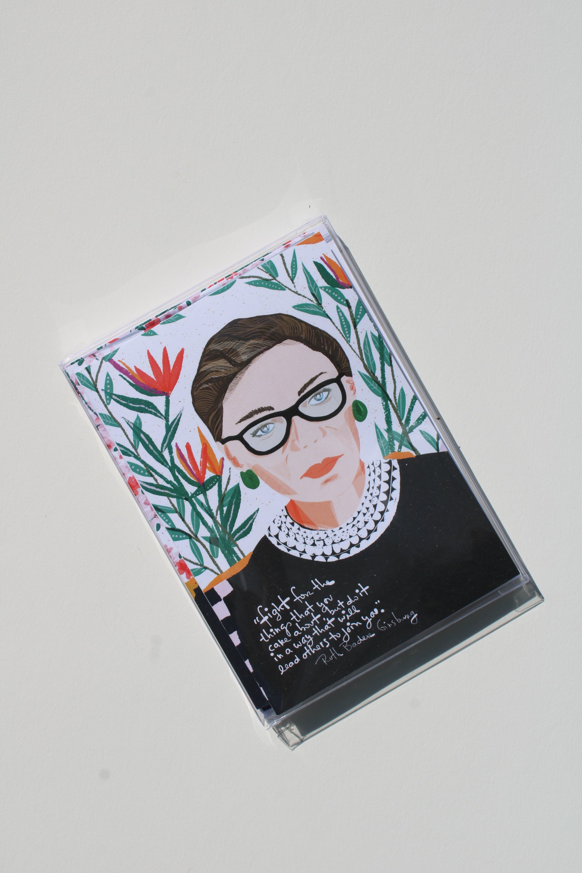 A clear plastic cassette tape case featuring a stylized portrait of Ruth Bader Ginsburg, surrounded by colorful tropical floral illustrations and handwritten text, shown on a light gray background.