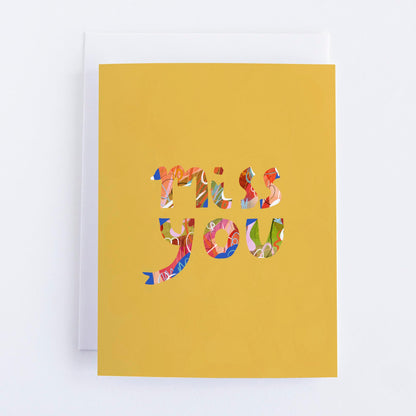 A yellow greeting card with the words 'Miss You' cut out in a colorful pattern from the card's material.